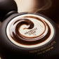 Angle : Gros plan macro serré, angle à $45^\circ$ centré sur le médaillon blanc et le tourbillon.Scène : Focus absolu sur le flacon de "French Vanilla Latte" de l'image image_8890cf.jpg. L'éclairage de studio (boîte à lumière focalisée) fait briller intensément le tourbillon marron au centre, créant des reflets spéculaires parfaits qui contrastent avec le fini mat marron foncé du reste du flacon. Une infime volute de vapeur chaude passe en arrière-plan.Fond : Arrière-plan flou de studio (bokeh) dégradé dans