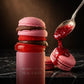 Angle : Vue macro de profil, focus sur le bouchon en forme de macarons empilés.  Scène : Le capuchon sculptural de l'image image_8c159e.png est mis en avant. Juste à côté de lui, une cuillère en argent vintage laisse couler un coulis de fruits rouges épais, brillant et texturé qui nappe artistiquement un vrai macaron croustillant posé sur le côté. Une pincée de poudre de cacao fine flotte subtilement dans l'air.  Décor & Lumière : Table de chef en marbre noir veiné. Lumière chaude et focalisée (style clair-