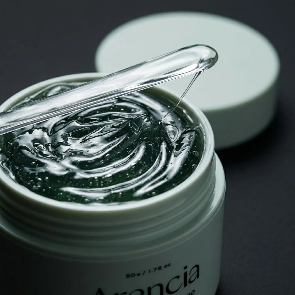 Angle : Gros plan macro très serré, focus sur la surface du gel ondulé.  Scène : Le pot d'Arencia de l'image image_c21e9d.jpg est ouvert, révélant la texture gélifiée et brillante du sérum vert forêt. Le couvercle mat céladon est posé en arrière-plan, légèrement flou. Une spatule en verre transparent est délicatement posée sur le rebord du pot, un fil de sérum brillant s'étirant depuis sa pointe.  Fond & Lumière : Arrière-plan sombre et minimaliste. Un éclairage rasant de studio fait miroiter les ondulation
