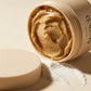 Angle : Gros plan macro très serré, focus sur le filetage du pot et la pâte.  Scène : Le pot d'Arencia est ouvert, révélant la texture compacte et élastique de sa pâte couleur ocre-miel. Le couvercle mat beige est posé à plat au premier plan, légèrement flou. Une fine traînée de poudre de riz blanc est artistiquement étalée à côté, contrastant avec l'onctuosité de la texture mochi.  Fond & Lumière : Arrière-plan épuré couleur crème. Un éclairage rasant de studio met en relief chaque relief et ondulation scu