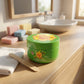 A commercial three-quarter photograph (angle de trois quarts) set on a light wood vanity in a stylized bathroom. The 200ml Chupa Chups - Apple Crush - Whipped Soap pot is positioned centrally, viewed from a slightly high front-right angle. A flat wooden stick is placed next to it, resting on a white towel. Several unbranded, pastel-colored bar soaps are arranged decoratively in the background, near a porcelain sink and a mirror. Warm sunlight streams in, creating soft shadows (gemini-3.1-flash-image-preview