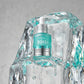 Angle : Contre-plongée légère (Low-angle).  Scène : Le flacon Anua est majestueusement posé sur un bloc de glace sculpté, parfaitement transparent et semi-fondu. L'eau qui fond crée des lignes fluides le long de la glace. La lumière traverse le bloc et le flacon en créant des reflets prismatiques turquoise et argentés éclatants.  Fond : Un arrière-plan minimaliste gris perle texturé de style studio éditorial. (gemini-3-pro-image-preview)