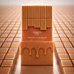 Une vue d'artiste carrée à 1200x1200px. La bouteille d'image_1.png est centrée, mais entourée d'une grille de carrés de chocolat au caramel identiques, créant un motif répétitif infini qui s'étire vers un horizon flou. La bouteille centrale est la seule parfaitement nette et colorée, les autres étant légèrement délavées. Une source de lumière frontale forte fait briller les arrêtes de la bouteille, la faisant ressortir comme un joyau dans la masse. (gpt-image-1.5)