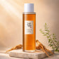 Creative e-commerce shot of the Beauty of Joseon Ginseng Essence Water bottle inspired by image_464141.png. The transparent bottle rests elegantly on a raw light beige stone platform, surrounded by authentic dried ginseng roots and a delicate sprig of green botanical leaves. The background is a soft, blurry warm ivory and amber-gold gradient under gentle morning sunbeams. Revitalizing, organic, and luxury Korean skincare concept. Aspect ratio 1:1, size 1200x1200px. (gpt-image-1.5)