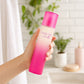 Scène : Photographie de style lifestyle. Une main féminine soignée tient délicatement le flacon "Simply Pink by Pink Sugar", s'apprêtant à l'utiliser après une douche. La main est positionnée naturellement. La lumière est douce et flatteuse.  Angle : Prise de vue à niveau, légèrement oblique.  Fond : Une salle de bain minimaliste floue, avec des carreaux de métro blancs et des plantes vertes. (gpt-image-2)